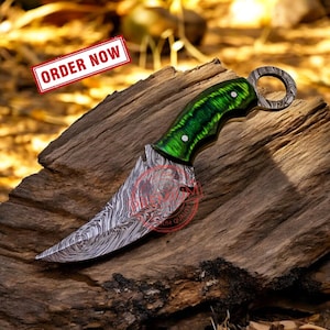 May include: A green and silver karambit knife with a ring handle. The blade is curved and has a Damascus steel pattern. The handle is made of wood and has a green epoxy resin inlay.