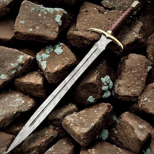 May include: A long, ornate sword with a patterned blade and a brown leather-wrapped handle. The sword features a gold-colored crossguard and pommel. The blade is approximately 30 inches long, and the sword rests on a bed of brown rocks.