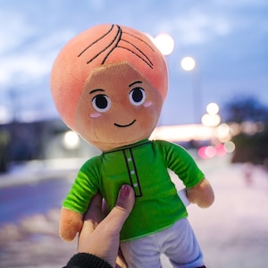 May include: A plush toy of a person with pink hair, a green shirt, and white pants. The toy is being held by a hand.