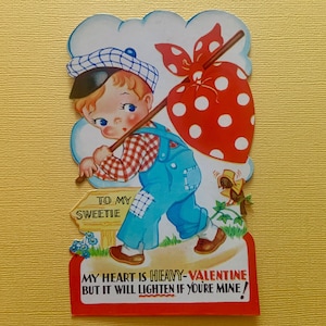 May include: A vintage Valentine's Day card featuring a cartoon boy carrying a red heart-shaped bag with white polka dots. The boy is wearing a blue overall, a red and white checkered shirt, and a blue and white baseball cap. The card has the text "To my sweetie" and "My heart is heavy-Valentine but it will lighten if you're mine!"