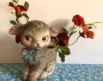 May include: A ceramic planter shaped like a lamb with a blue bow. The planter is filled with artificial red flowers.