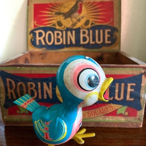 May include: A vintage tin wind-up bird toy painted in bright blue, pink, and yellow, featuring large cartoon eyes and an open beak, positioned in front of an open wooden box labeled with the text ROBIN BLUE and an illustration of a robin.