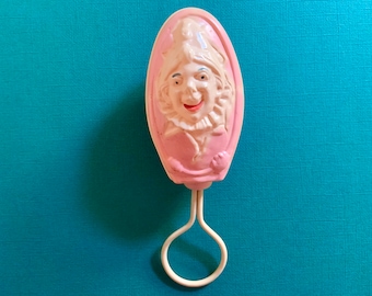 May include: A pink plastic baby rattle with a white handle. The rattle features a smiling face with a white hat and a pink ruffle.