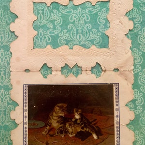 May include: A vintage greeting card featuring a decorative white frame and a picture of a mother cat and two kittens. The kittens are brown and white, and the mother cat is brown and black. The picture is set against a dark background.