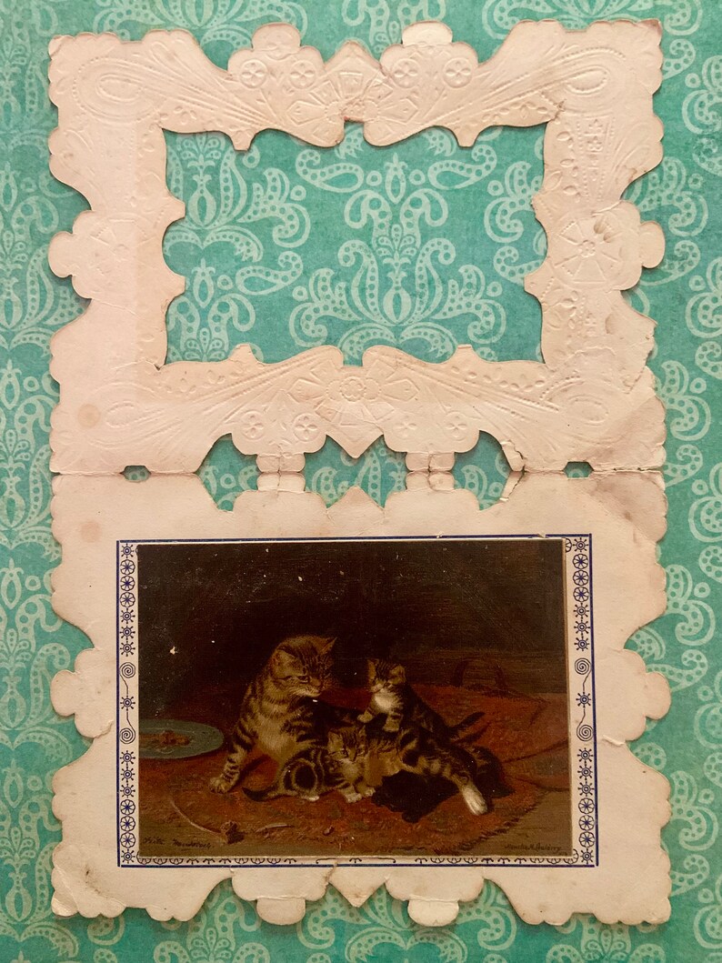 May include: A vintage greeting card featuring a decorative white frame and a picture of a mother cat and two kittens. The kittens are brown and white, and the mother cat is brown and black. The picture is set against a dark background.