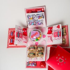 May include: An open, multi-layered gift box filled with various candies and chocolates. The box is decorated with pink hearts and red accents. A clear container with a heart-shaped treat and the letters "xoxo" is in the center. Several miniature candy bars are also visible.