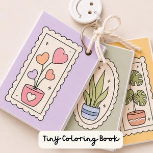 May include: A set of three small rectangular coloring books bound together by a metal ring and a white smiley face charm. The covers feature illustrations of potted plants in scalloped frames with pastel purple, green, and yellow backgrounds.