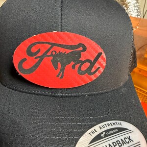 May include: Black snapback hat with a red oval patch featuring the Ford logo and a horse silhouette. The hat has a mesh back and a curved brim. The tag reads "Snapback".