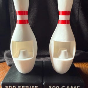 May include: Two white bowling pins with red stripes, each on a black base. The bases read "800 SERIES" and "300 GAME" in white text. The pins have a hollowed-out design.