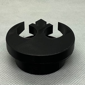 May include: A black plastic 3D printed object shaped like the Rebel Alliance symbol from Star Wars. The symbol is a circle with a crescent moon shape on the left side and a triangle shape on the right side. The object is a bottle stopper.