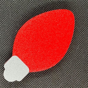 May include: A red Christmas light bulb ornament with a silver base. The ornament is a solid, textured red color and is set against a black background with white polka dots. The bulb shape is elongated.