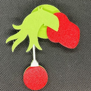 May include: Green felt Grinch hand holding a red felt Christmas ornament. The ornament is attached to the hand with a white felt stem.