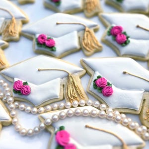 May include: Graduation-themed cookies shaped like white mortarboards with gold tassels and pink rose decorations. A string of pearls is draped across the cookies, creating a celebratory and elegant display.