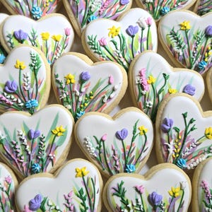 May include: A collection of heart-shaped sugar biscuits decorated with white icing and hand-painted floral designs. Each biscuit features colourful icing accents in shades of yellow, purple, pink, and green, depicting various wildflowers and stems.