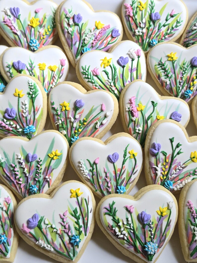 May include: A collection of heart-shaped sugar biscuits decorated with white icing and hand-painted floral designs. Each biscuit features colourful icing accents in shades of yellow, purple, pink, and green, depicting various wildflowers and stems.