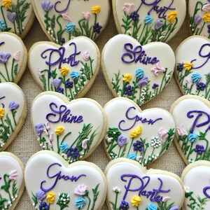May include: Heart-shaped sugar biscuits with white icing, decorated with hand-piped purple script text reading Grow, Plant, or Shine, and colourful floral designs featuring green stems, yellow, blue, pink, and purple icing flowers.