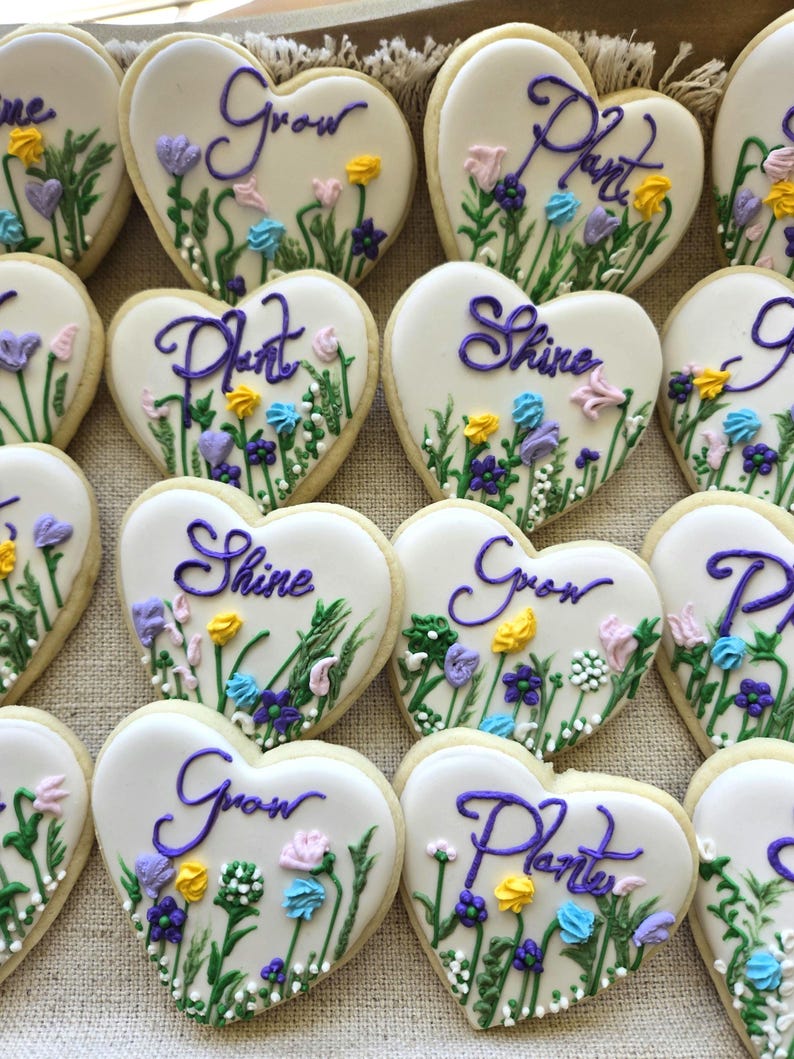 May include: Heart-shaped sugar biscuits with white icing, decorated with hand-piped purple script text reading Grow, Plant, or Shine, and colourful floral designs featuring green stems, yellow, blue, pink, and purple icing flowers.