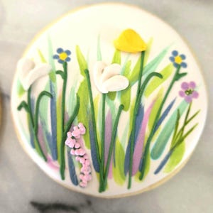 May include: A round sugar biscuit decorated with white icing featuring a hand-painted meadow scene. The design includes green grass blades, small blue and purple flowers, a single yellow petal, and white dimensional icing blossoms.