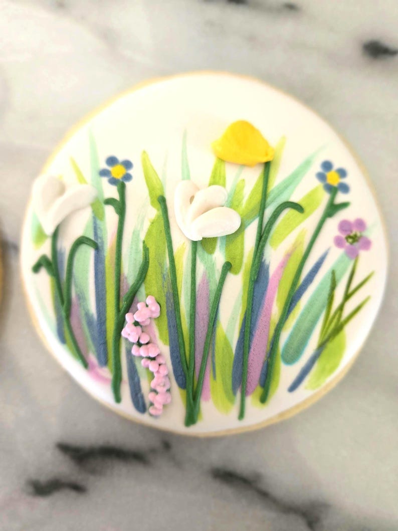 May include: A round sugar biscuit decorated with white icing featuring a hand-painted meadow scene. The design includes green grass blades, small blue and purple flowers, a single yellow petal, and white dimensional icing blossoms.