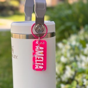 K&ouml;nnte beinhalten: Ein wei&szlig;er Stanley-Becher mit grauem Griff und einem rosa Schl&uuml;sselanh&auml;nger. Der Schl&uuml;sselanh&auml;nger ist rechteckig und tr&auml;gt den Namen "AMELIA" in wei&szlig;en Buchstaben. Der Anh&auml;nger hat auch ein florales Design.