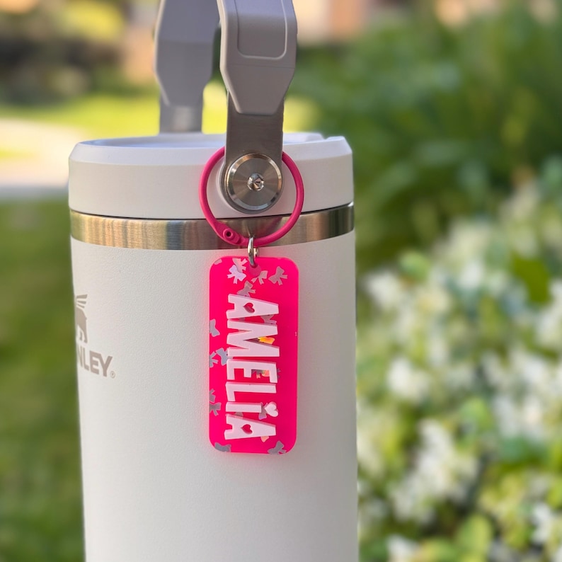 K&ouml;nnte beinhalten: Ein wei&szlig;er Stanley-Becher mit grauem Griff und einem rosa Schl&uuml;sselanh&auml;nger. Der Schl&uuml;sselanh&auml;nger ist rechteckig und tr&auml;gt den Namen "AMELIA" in wei&szlig;en Buchstaben. Der Anh&auml;nger hat auch ein florales Design.