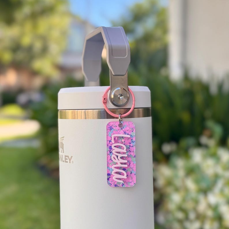 K&ouml;nnte beinhalten: Eine wei&szlig;e, isolierte Wasserflasche mit grauem Griff und einem rosa Schl&uuml;sselanh&auml;nger. Der Schl&uuml;sselanh&auml;nger hat ein rechteckiges Etikett mit dem Namen "Laya" in wei&szlig;er Schrift auf einem rosa-lila Glitzerhintergrund.