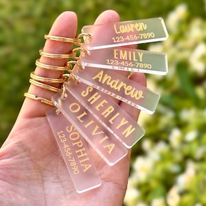 May include: A hand holds a collection of personalized keychains. Each rectangular keychain is clear with gold lettering, displaying names and phone numbers. The keychains are attached to gold-colored key rings. The background is a soft, blurred green.