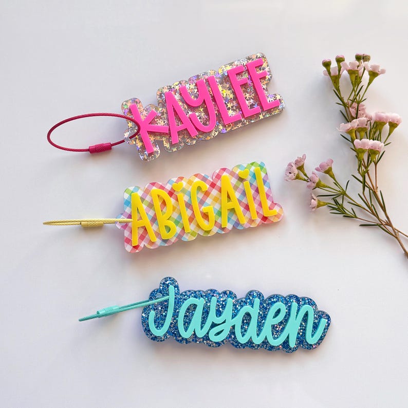 May include: Three personalized name tags in a row. The top tag is pink with the name "Kaylee" in a bubble font. The middle tag has the name "Abigail" in yellow. The bottom tag is blue with the name "Jayden" in a script font.