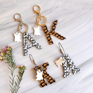 May include: Halloween-themed keychains featuring the letters "A" and "K". The letters are decorated with either a black and white ghost pattern or a black and orange pumpkin pattern. Each keychain includes a small white ghost charm and a metal clasp.