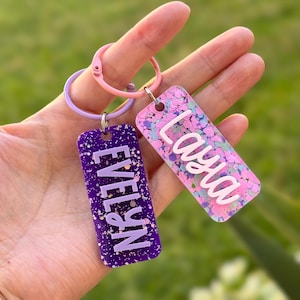 K&ouml;nnte beinhalten: Zwei rechteckige Schl&uuml;sselanh&auml;nger mit Namen. Einer ist lila mit dem Namen "Evelyn" in Wei&szlig;, der andere ist rosa mit dem Namen "Laya" in Wei&szlig;. Beide Schl&uuml;sselanh&auml;nger haben Glitzerakzente und sind an einem rosa und lila Schl&uuml;sselring befestigt.