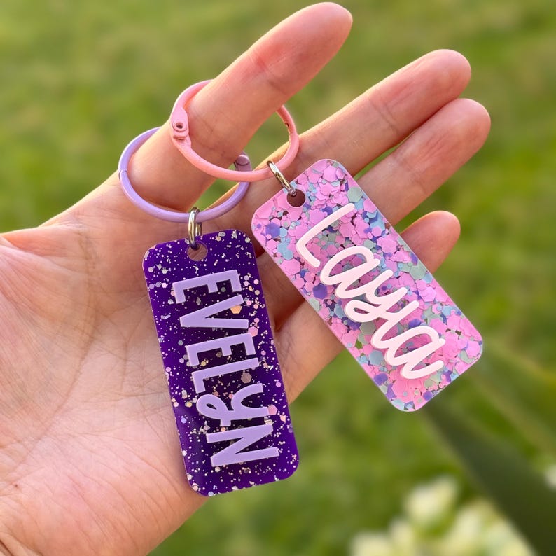 K&ouml;nnte beinhalten: Zwei rechteckige Schl&uuml;sselanh&auml;nger mit Namen. Einer ist lila mit dem Namen "Evelyn" in Wei&szlig;, der andere ist rosa mit dem Namen "Laya" in Wei&szlig;. Beide Schl&uuml;sselanh&auml;nger haben Glitzerakzente und sind an einem rosa und lila Schl&uuml;sselring befestigt.