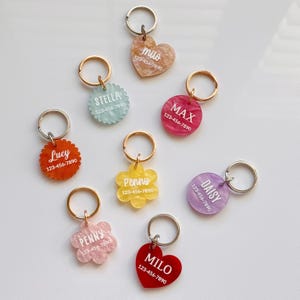 Puede incluir: Una colecci&oacute;n de etiquetas de identificaci&oacute;n para mascotas coloridas en varias formas, incluyendo corazones, flores y c&iacute;rculos. Cada etiqueta presenta un nombre y un n&uacute;mero de tel&eacute;fono en texto blanco, adjunta a un llavero de metal. Los colores son rojo, rosa, amarillo, azul y morado.