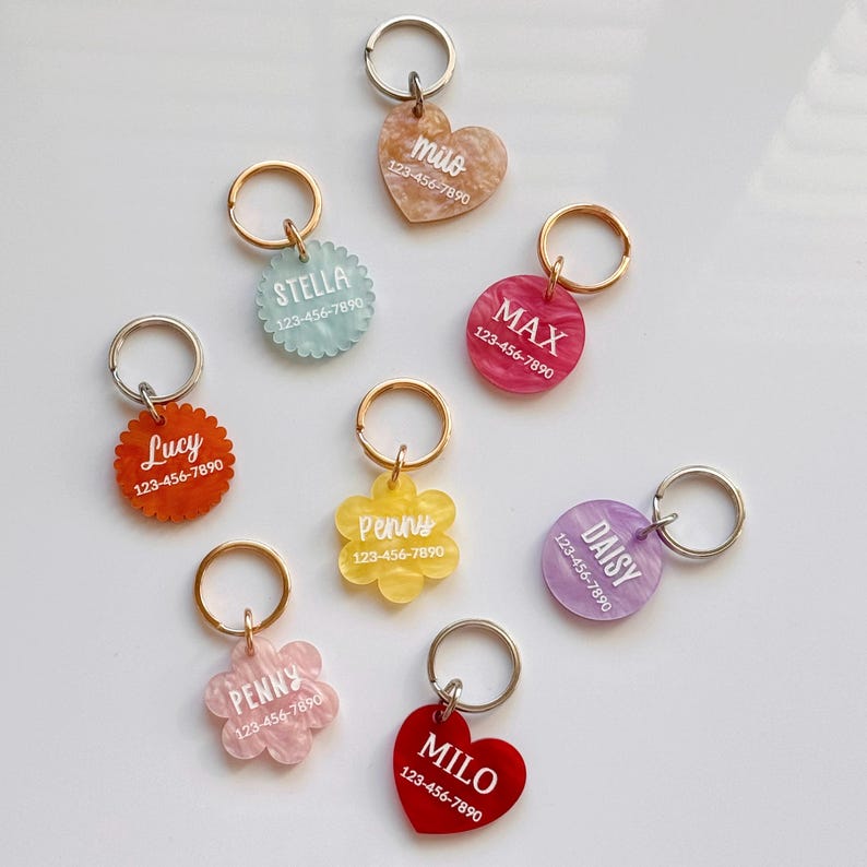 Puede incluir: Una colecci&oacute;n de etiquetas de identificaci&oacute;n para mascotas coloridas en varias formas, incluyendo corazones, flores y c&iacute;rculos. Cada etiqueta presenta un nombre y un n&uacute;mero de tel&eacute;fono en texto blanco, adjunta a un llavero de metal. Los colores son rojo, rosa, amarillo, azul y morado.