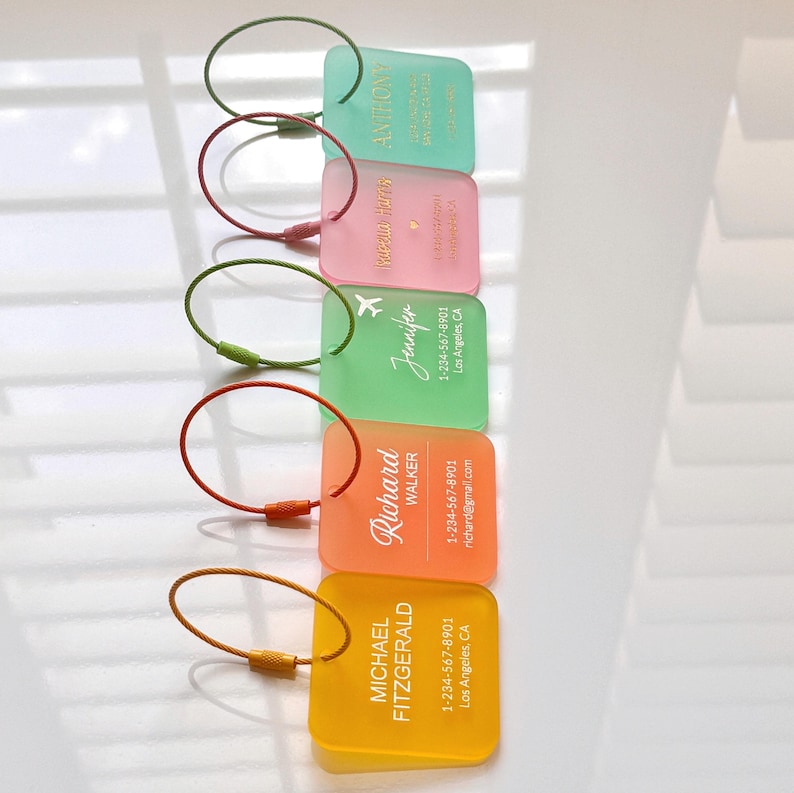 May include: A stack of colorful luggage tags in shades of green, pink, orange, and yellow. Each tag has a metal loop and features a name and contact information. The tags are rectangular with rounded corners.