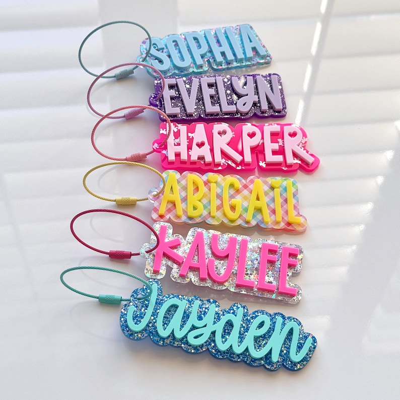 May include: A collection of personalized name tags in various colors. Each tag features a different name: Sophia, Evelyn, Harper, Abigail, Kaylee, and Jayden. The tags are made of glittery acrylic and have a metal loop for attachment.