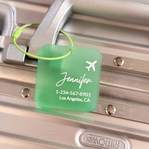 May include: A green luggage tag with a white airplane icon and the name "Jennifer" in script. Below the name is a phone number and the city and state, Los Angeles, CA. The tag is attached to a silver suitcase handle with a green loop.