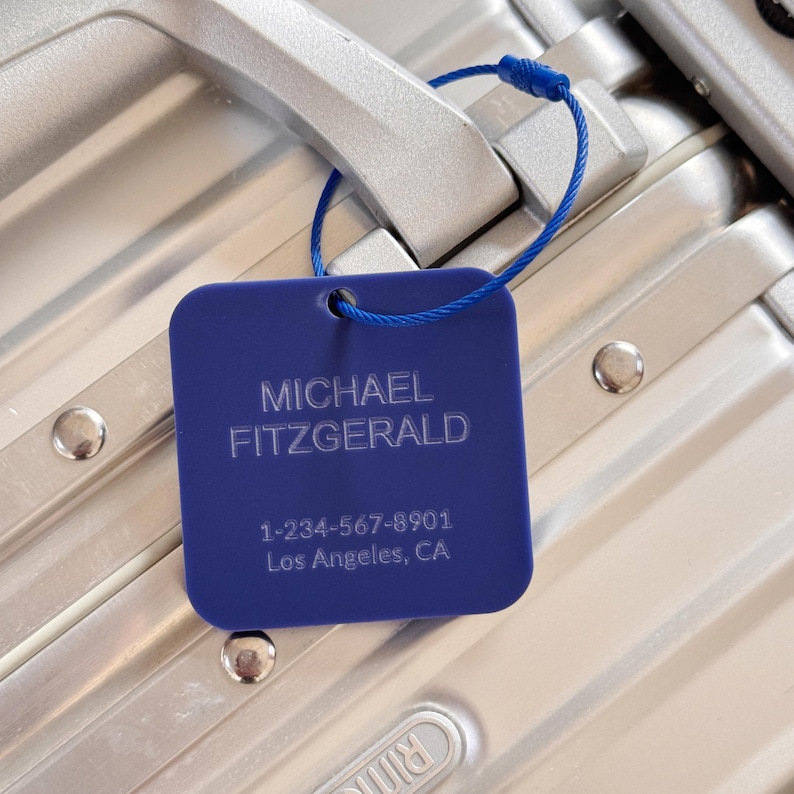 May include: A blue luggage tag with the name "MICHAEL FITZGERALD", a phone number, and the city "Los Angeles, CA" printed in white. The tag is attached to a silver suitcase.