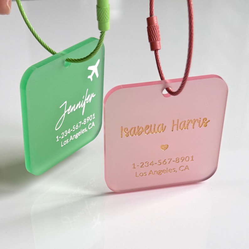 May include: Two personalized luggage tags. One is green with white text and a small airplane graphic. The other is pink with gold text and a heart graphic. Both tags have a phone number and city.