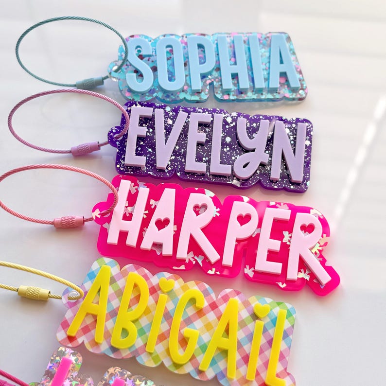 May include: Four personalized name tags in various colors. The names are "Sophia", "Evelyn", "Harper", and "Abigail". Each tag has a different color scheme, including blue, purple, pink, and yellow. The tags are attached to a metal loop.