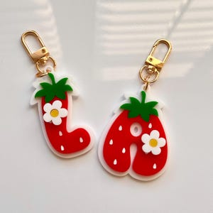 May include: Two strawberry-shaped keychains. The keychains are red with white seeds and a white outline. Each has a green leafy top and a small white flower with a yellow center. Gold-colored clasps are attached.