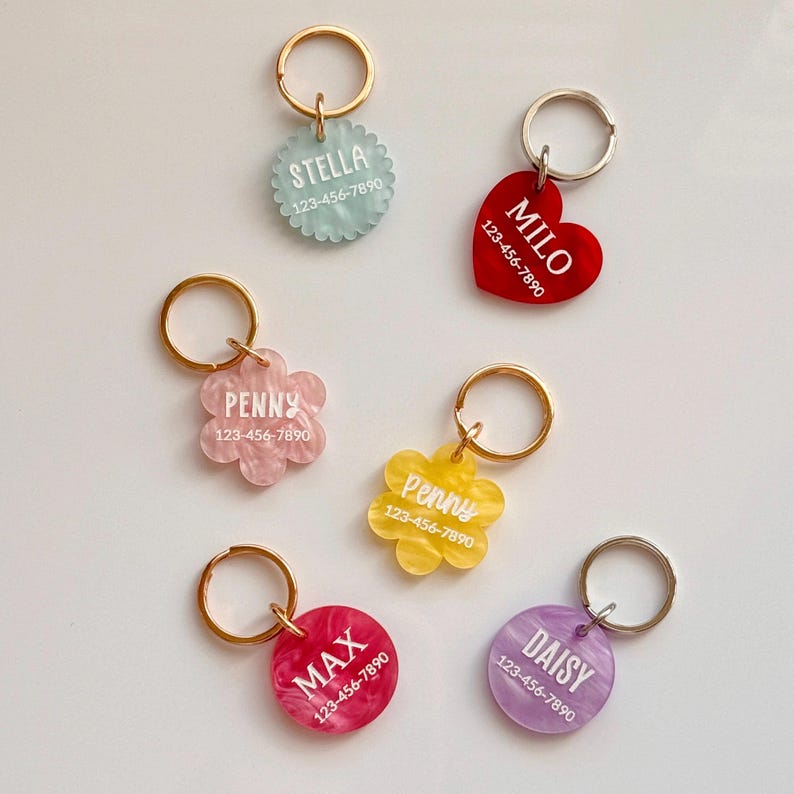 Puede incluir: Una colecci&oacute;n de etiquetas de identificaci&oacute;n para mascotas de colores en varias formas, incluyendo un coraz&oacute;n, flores y c&iacute;rculos. Cada etiqueta presenta un nombre y un n&uacute;mero de tel&eacute;fono en texto blanco, adjunto a un llavero dorado o plateado. Colores: rojo, rosa, amarillo, morado y turquesa.