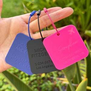 May include: Three square luggage tags in blue, black, and hot pink, each featuring a metal cable loop. The tags are engraved with names, phone numbers, and city locations. The pink tag displays the name Seraphina and a small airplane icon.