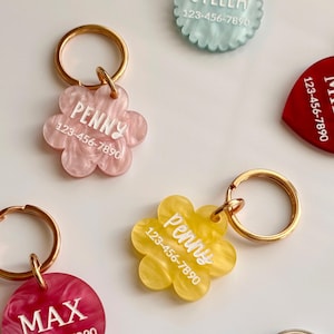 Puede incluir: Una colecci&oacute;n de etiquetas de identificaci&oacute;n para mascotas en varios colores y formas. Las etiquetas tienen forma de flor, coraz&oacute;n y circular, con llaveros dorados. Cada etiqueta presenta el nombre de una mascota y un n&uacute;mero de tel&eacute;fono en letras blancas.