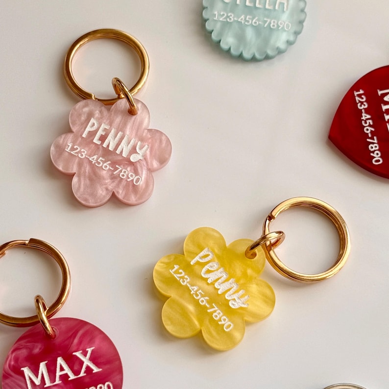 Puede incluir: Una colecci&oacute;n de etiquetas de identificaci&oacute;n para mascotas en varios colores y formas. Las etiquetas tienen forma de flor, coraz&oacute;n y circular, con llaveros dorados. Cada etiqueta presenta el nombre de una mascota y un n&uacute;mero de tel&eacute;fono en letras blancas.