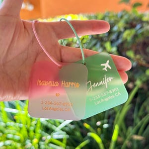 May include: Two personalized luggage tags. One tag is pink and green with the name "Isabella Harris" in gold lettering. The other tag is green with the name "Jennifer" and a small airplane icon. Both tags have contact information.