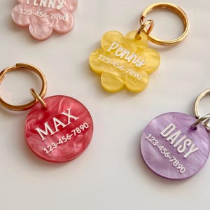 Puede incluir: Primer plano de varias etiquetas de identificaci&oacute;n para mascotas en varios colores y formas. Las etiquetas tienen forma de flores y c&iacute;rculos. Cada etiqueta tiene un nombre y un n&uacute;mero de tel&eacute;fono impresos. Las etiquetas est&aacute;n unidas a llaveros dorados.