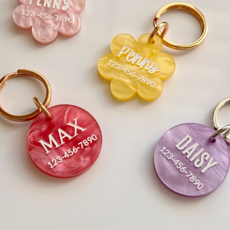 Puede incluir: Primer plano de varias etiquetas de identificaci&oacute;n para mascotas en varios colores y formas. Las etiquetas tienen forma de flores y c&iacute;rculos. Cada etiqueta tiene un nombre y un n&uacute;mero de tel&eacute;fono impresos. Las etiquetas est&aacute;n unidas a llaveros dorados.