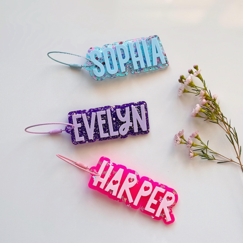 May include: Three personalized name tags in blue, purple, and pink. The tags are rectangular with rounded edges and feature names in white lettering. Each tag has a matching colored loop for attachment.