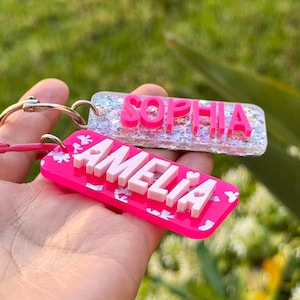 K&ouml;nnte beinhalten: Zwei personalisierte Schl&uuml;sselanh&auml;nger. Einer ist pinkfarben mit dem Namen "AMELIA" in wei&szlig;en Buchstaben, und der andere ist transparent mit dem Namen "SOPHIA" in pinkfarbenen Buchstaben. Beide Schl&uuml;sselanh&auml;nger haben silberne Schl&uuml;sselringe und werden in einer Hand gehalten.