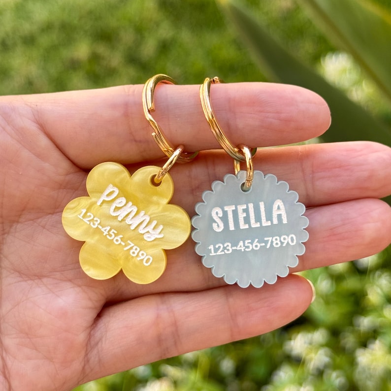 Puede incluir: Dos etiquetas de identificaci&oacute;n para mascotas con llaveros dorados. Una es amarilla, con forma de flor, y dice "Penny" y un n&uacute;mero de tel&eacute;fono. La otra es azul claro, festoneada, y dice "Stella" y un n&uacute;mero de tel&eacute;fono.